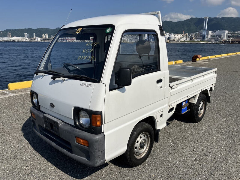 White 1990 Subaru Sambar KS4 Kei Truck with AWD, parked by the waterfront, featuring square headlights, robust front bumper, and open cargo bed, highlighted on Oiwa Garage’s Kei truck information page.