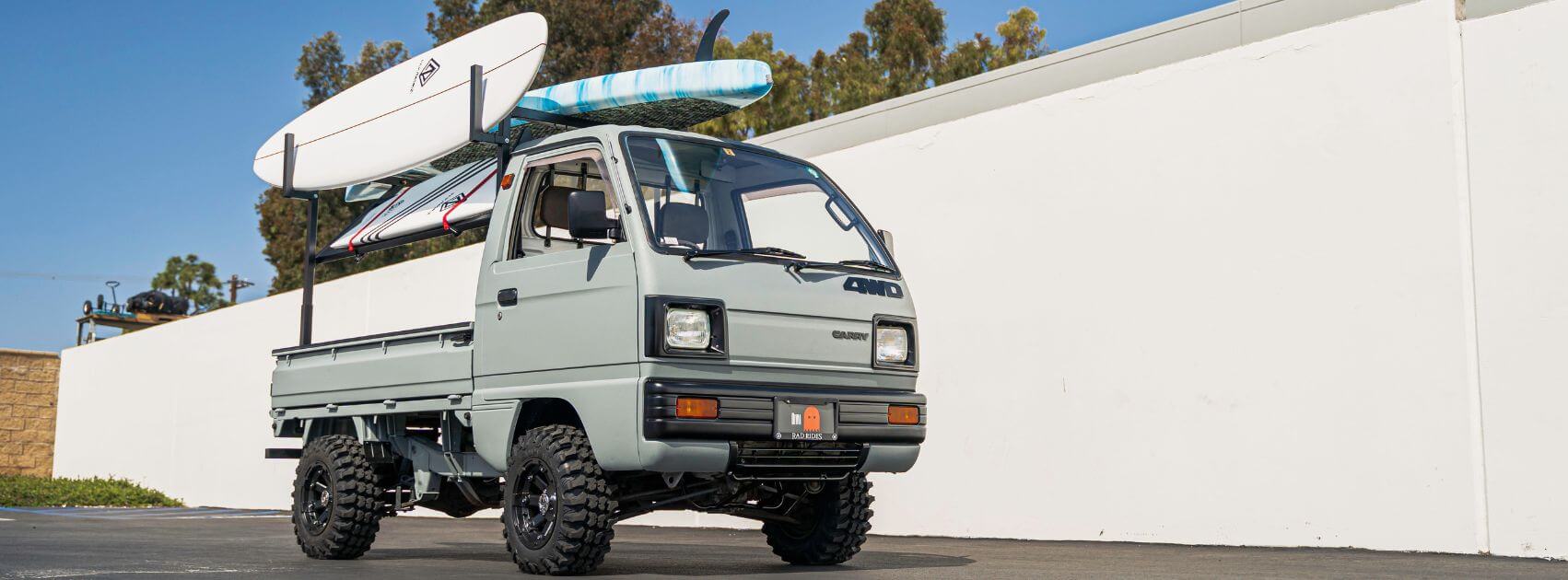 Eighth Generation Suzuki Carry DB71T with Surfboards, featuring upgraded suspension and off-road tires, parked beside a white wall, showcasing its robust design and cargo versatility perfect for outdoor adventures.