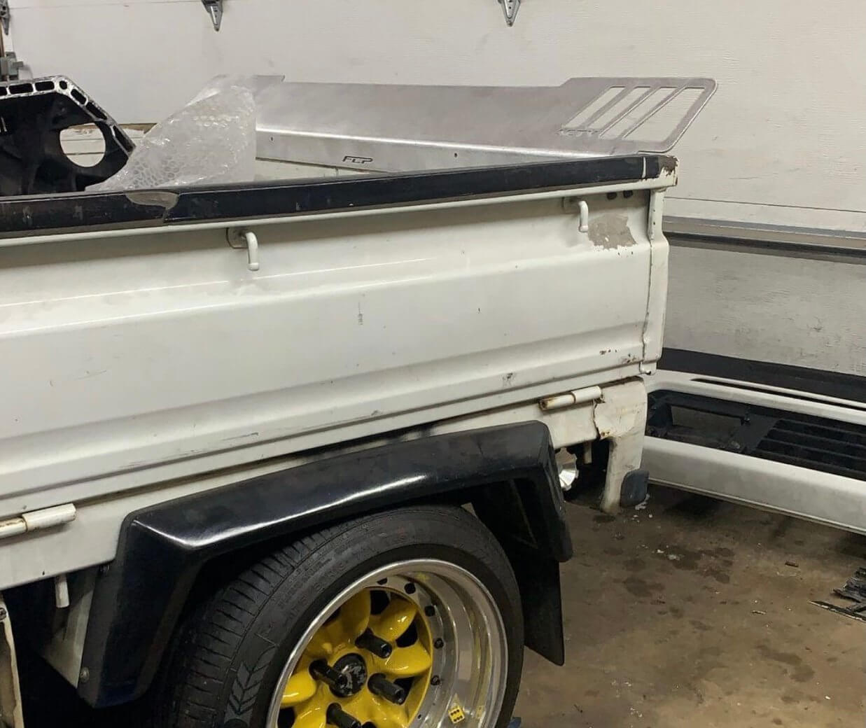 Rear Lusso Spoiler for Honda Acty Truck, silver aluminum, mounted on a white truck bed, with black wheel and garage background