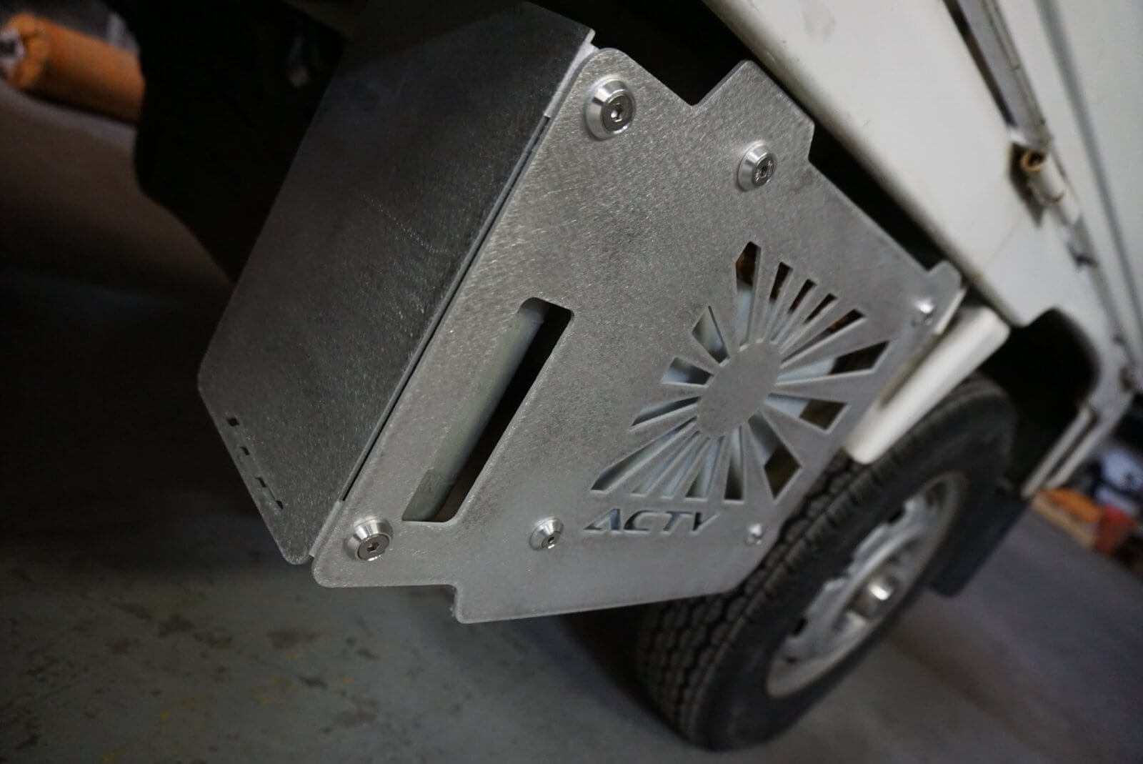 Rising Sun Battery Cover for Honda Acty Truck, silver aluminum with sun design, mounted at an angle, industrial background