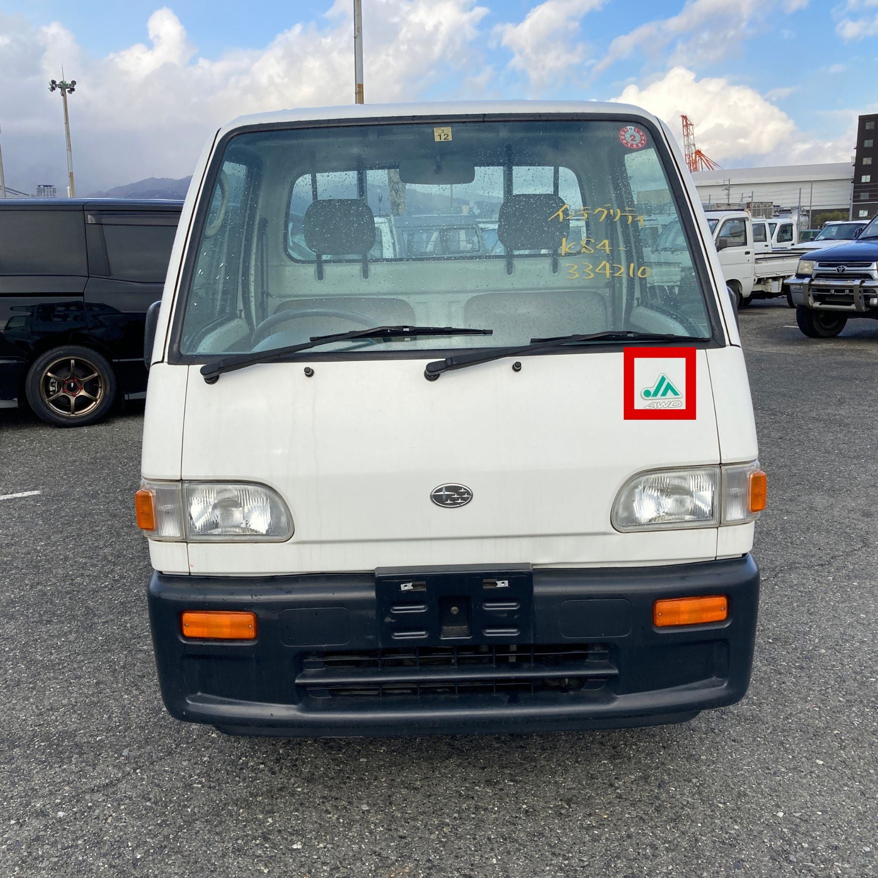 Front view of a white Subaru Sambar KS3/KS4 kei truck parked in a lot. A green JA decal, highlighted in red, is placed on the right side of the truck's front panel above the headlight. The windshield has yellow writing with vehicle information.