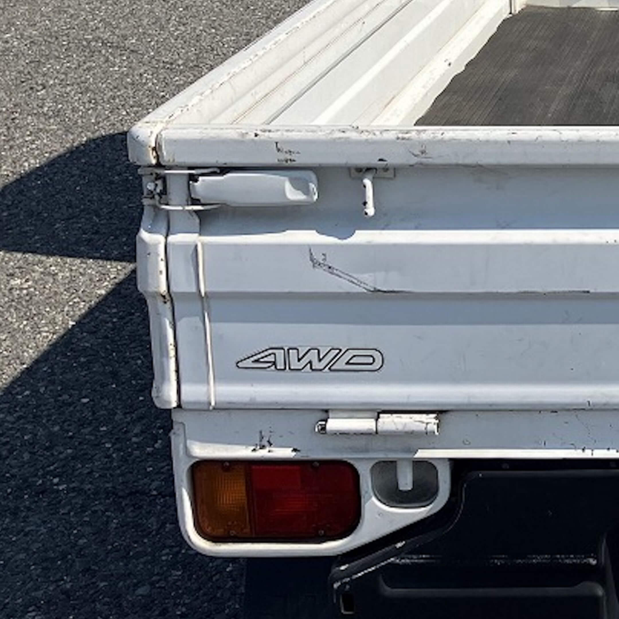 Close-up of the rear left side of a white Subaru Sambar KS3/KS4 truck tailgate. The tailgate features a black "4WD" decal. Below the decal, the corner taillight with amber and red lenses is visible.
