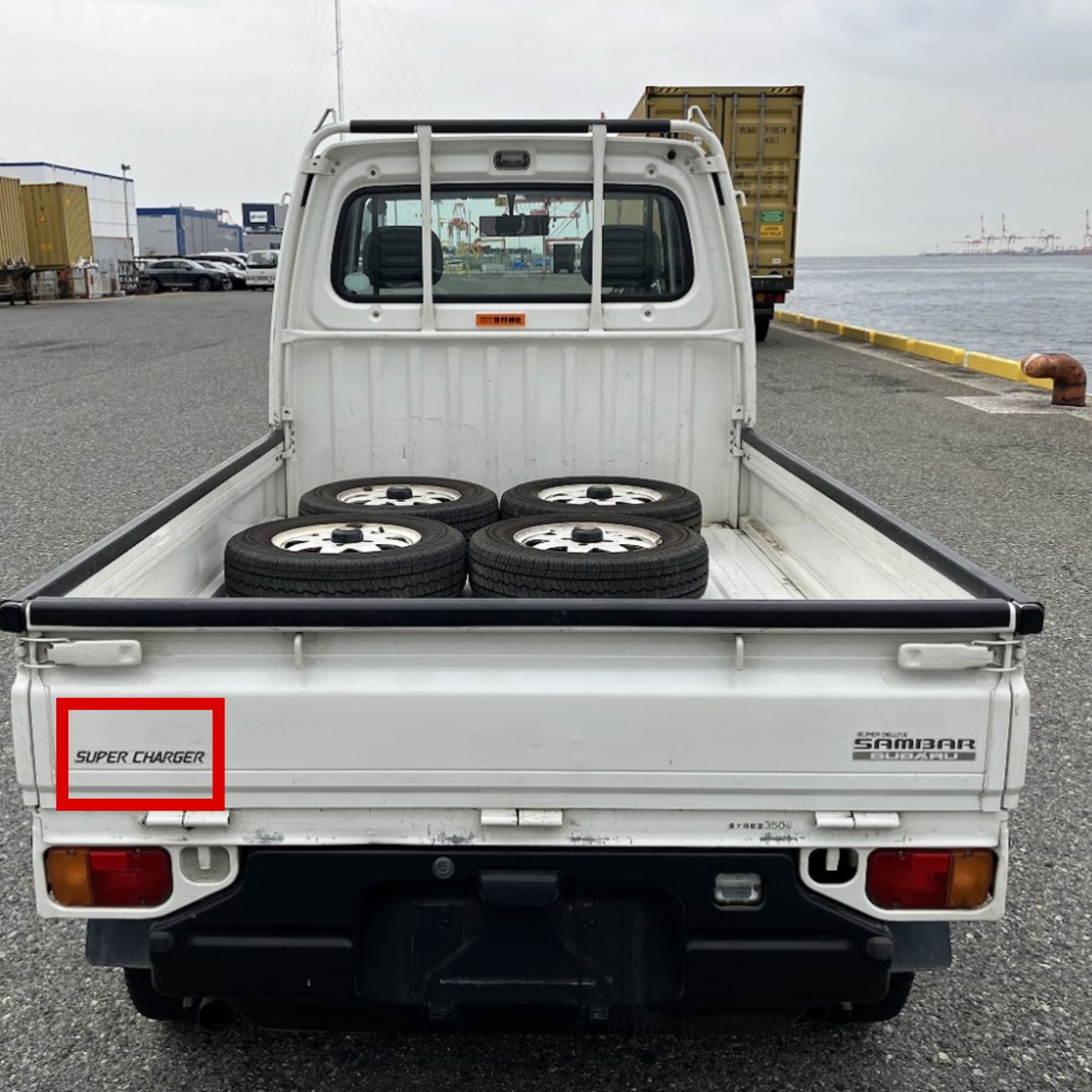 Rear view of a white Subaru Sambar kei truck with its tailgate closed. The bed contains four spare tires. A "Super Charger" decal, highlighted in red, is visible on the left side of the tailgate, while "Sambar Subaru" branding is on the right side